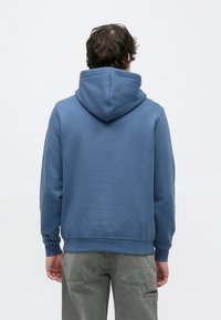 Μπλε φούτερ με κουκούλα και κορδόνι, υφασμάτινη υφή, ριμπ μανσέτες και μέση. Φορεμένο με ανοιχτό γκρι παντελόνι cargo.