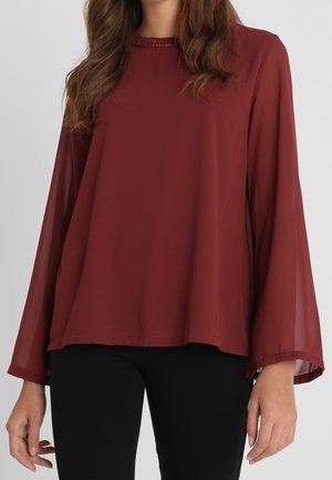 Woman wearing a loose, long-sleeve, dark red blouse with subtle neckline detail and black pants, standing against a plain background.