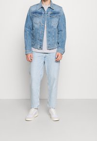 Ljusblå denimjacka med klassisk design, med knappstängning och bröstfickor, bärs över en grå t-shirt och ljusblå jeans.