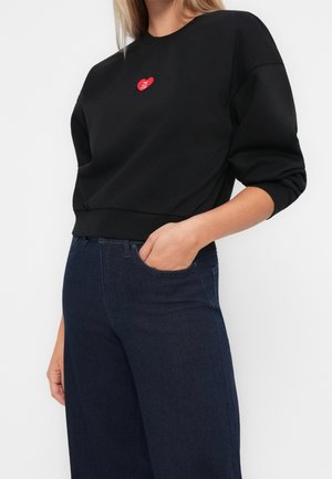 Femme portant un sweat-shirt noir avec un petit logo cœur rouge sur la poitrine et un jean bleu foncé taille haute, main dans la poche.