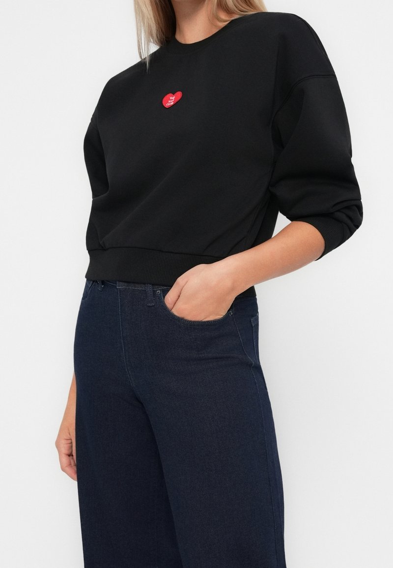 Femme portant un sweat-shirt noir avec un petit logo cœur rouge sur la poitrine et un jean bleu foncé taille haute, main dans la poche.