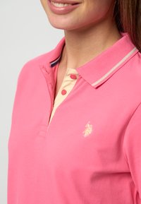 Posnetek od blizu nasmejane osebe, ki nosi roza polo majico z bež notranjo zaponko in majhnim vezenim logotipom na prsih.