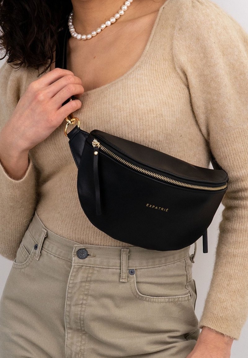 Černá kožená taška přes rameno s zakřiveným tvarem, zlatým zipem a logem značky. Popruhy jsou připevněny se zlatými kovovými doplňky.