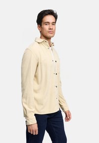 Camisa de pana beige con cuello doblado, botones en la parte delantera y un bolsillo en el pecho. Diseño ajustado con mangas largas y tela texturizada.