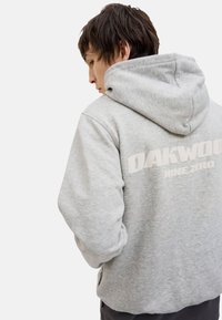 Hanorac gri din bumbac cu un mare logo alb "OAKWOOD" pe spate, dotat cu glugă cu șnur și manșete ribbate.