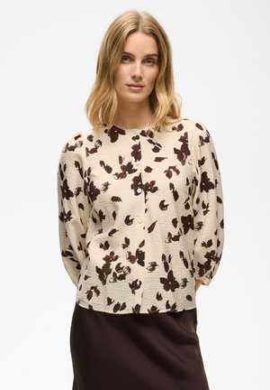 Kvinde iført en cremefarvet bluse med mørkebrune blomsterprint og et mørkebrunt skørt, stående mod en ensfarvet lys baggrund.