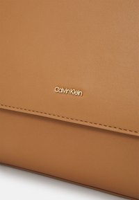 Béžová taška z umělé kůže s zlatým embosovaným logem "Calvin Klein". Hladký povrch, diskrétní švy podél okraje.