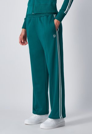 Pantaloni tuta turchesi con strisce laterali bianche, vita elastica e tasche laterali con zipper, abbinati a sneakers bianche. Texture del tessuto liscia.