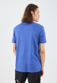 Tricou din bumbac albastru, cu mâneci scurte, guler rotund și design simplu. Cusături vizibile și o croială lejeră. Purtați cu blugi negri uzați.