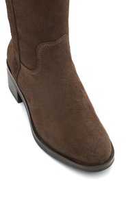 Brązowy zamszowy but za kostkę z okrągłym noskiem, widocznymi przeszyciami i drewnianym obcasem na stosie.