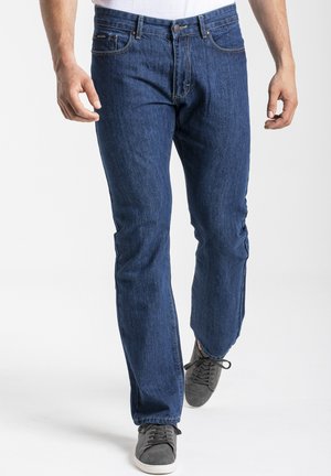 Jean en denim bleu avec une coupe droite, dotés de cinq poches, d'une fermeture éclair et de coutures contrastantes. Portés avec des baskets grises à lacets.