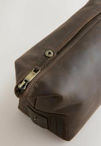 Punguță maro din piele cu corp texturat, închidere cu fermoar în partea de sus și un mâner metalic. Prezintă o bază dreptunghiulară și un design cusut pe o parte.