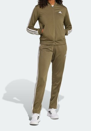 Person iført olivengrøn Adidas træningsdragt med hvide striber på ærmer og ben, hænderne i jakke-lommerne, stående på hvid baggrund.