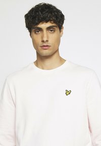 Felpa rosa chiaro realizzata in morbido tessuto, con scollo a girocollo e un piccolo logo giallo ricamato sulla parte sinistra del petto.