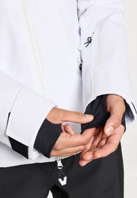 Bijela vodootporna jakna s crnim detaljima; ima prednji zip, podesive manžete i rupe za palčeve. Kratki pregled ruku koje podešavaju manžetnu.
