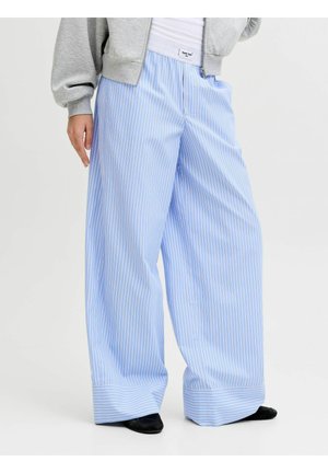 Blauw-witte broek met wijde pijpen, gedragen met een grijze jas met rits en zwarte schoenen, zichtbaar vanaf het midden van de torso naar beneden.