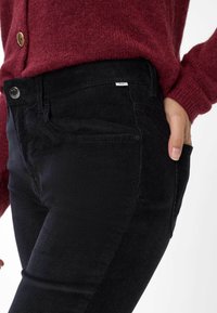 Schwarze Cordjeans mit strukturiertem Oberflächen und seitlichen Taschen, kombiniert mit einem burgunderfarbenen Strickpullover mit großen Knöpfen.