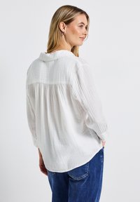 Kvinna med långt blont hår som bär en lös vit texturerad blus och blå jeans, står med händerna i fickorna och tittar åt sidan.