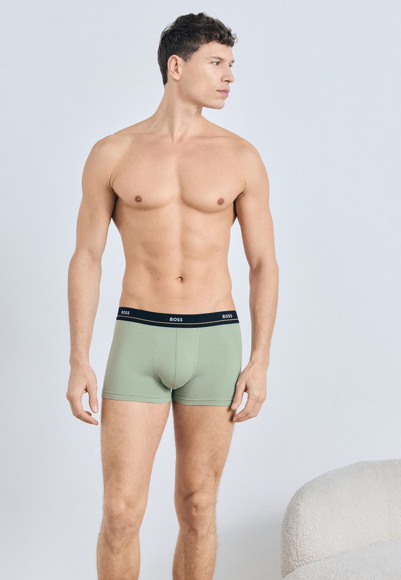 Zielone bokserki z czarnym pasem w talii, na którym znajduje się logo marki. Tkanina wydaje się być miękka i gładka. Krój jest dopasowany i o długości sięgającej do połowy uda.