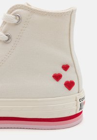 Vysoké plátenné tenisky v krémovej farbe s červenými vyšívanými srdiečkami. Obsahujú kovové očká, gumenú podrážku s červenými a ružovými akcentmi.