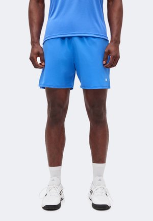 Uomo che indossa pantaloncini sportivi blu, calzini bianchi e scarpe da ginnastica bianche Adidas, in piedi su uno sfondo chiaro e uniforme.