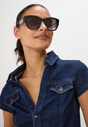 Donna con grandi occhiali da sole neri, una camicia di jeans blu con tasche e una collana d'oro con piccoli ciondoli, che guarda leggermente verso l'alto.