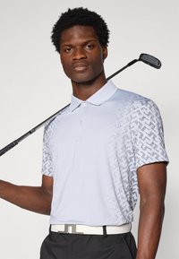 Svetlo modra polo majica s kratkimi rokavi, ki ima vzorčasto zasnovo na levem rami. Model drži golf palico, oblečen v črne hlače in belo pas.