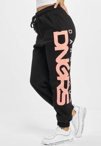 Black sweatpants with a wide pink "DNGRS" logo on the side, featuring an elastic waistband and cuffs, paired with white sneakers.