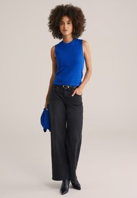 Un haut sans manches en tricot bleu associé à un jean noir à taille haute et jambes larges, porté avec des escarpins noirs à bout pointu,.accessoirisé d'un pull bleu assorti.
