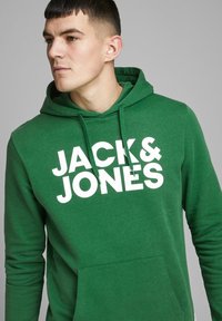 Felpa verde realizzata in tessuto morbido, con tasca a marsupio e un grande logo bianco "JACK & JONES" sul davanti. Inclusa cappuccio regolabile con coulisse.