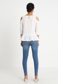 Blusa bianca con peplum, con tagli sulle spalle, tessuto strutturato, apertura a chiave sul retro e maniche a tre quarti, abbinata a jeans aderenti blu e tacchi.