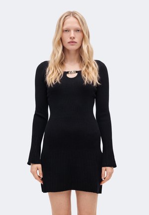 Femme aux longs cheveux blonds portant une robe en maille côtelée noire à manches longues avec une petite découpe et un détail boucle au niveau du col.
