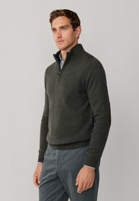 Donkergroene wollen trui met een ritskraag en geribde manchetten; bevat leren patches op de schouders en is gelaagd met een blauw geruit shirt.