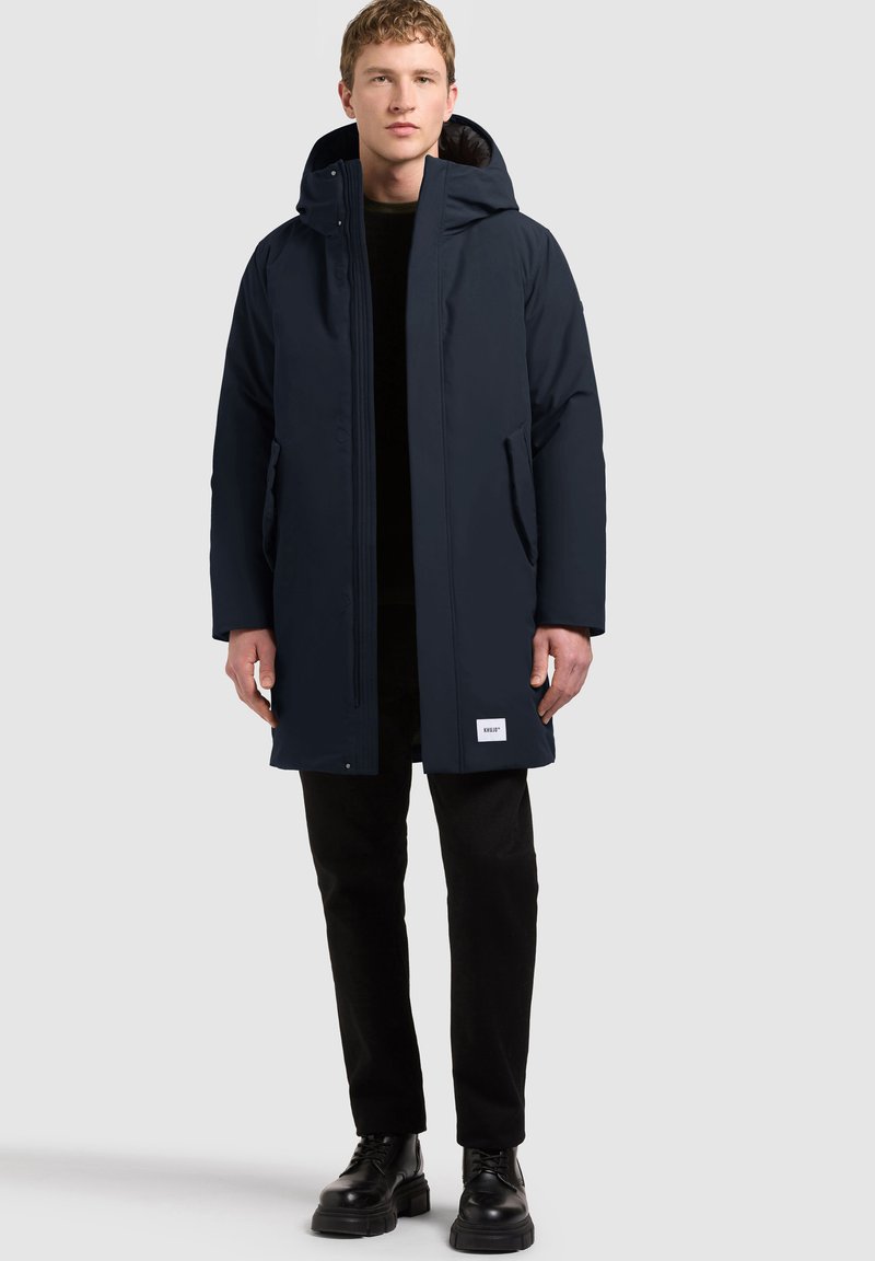 Jeune homme portant un long manteau d'hiver bleu foncé à capuche, un pantalon noir et des bottes noires à lacets, debout devant un fond clair uni.