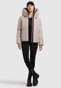 Hellgraue Steppjacke mit einer großen Kapuze, ausgestattet mit einem Reißverschluss vorne und zwei Seitentaschen, kombiniert mit schwarzen Leggings und Stiefeln.