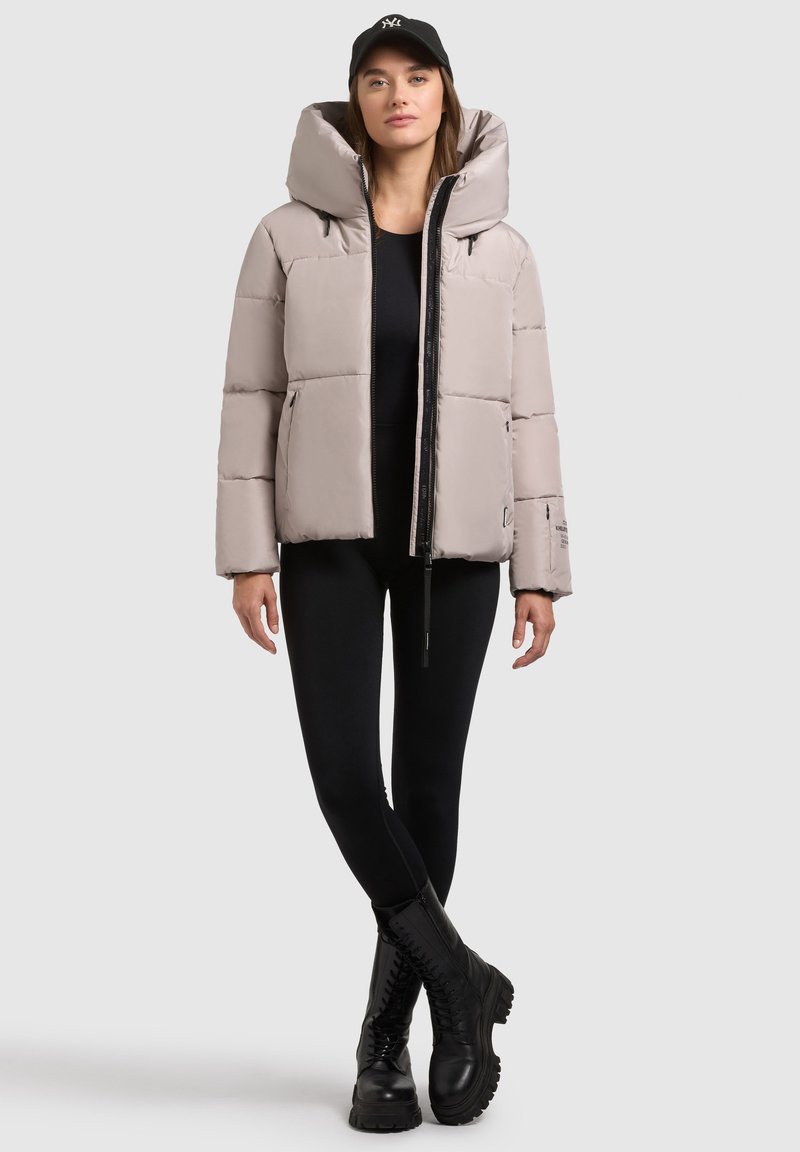 Hellgraue Steppjacke mit einer großen Kapuze, ausgestattet mit einem Reißverschluss vorne und zwei Seitentaschen, kombiniert mit schwarzen Leggings und Stiefeln.