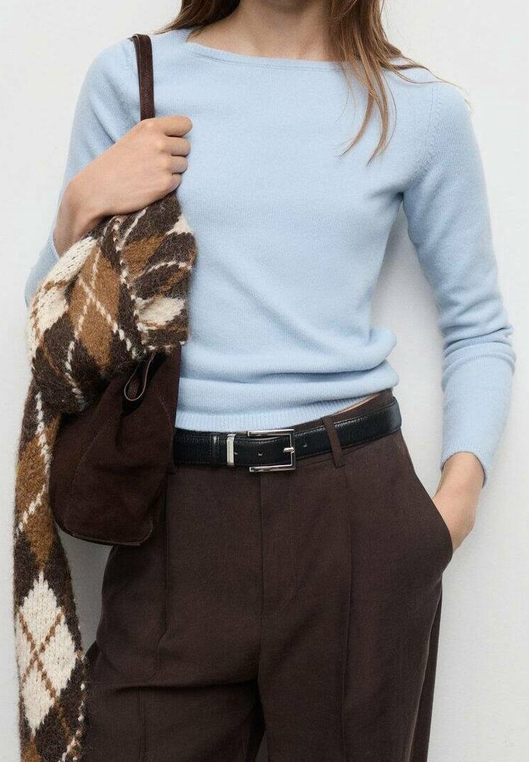 Femme vêtue d’un haut bleu clair à manches longues, d’un pantalon marron taille haute avec ceinture noire, tenant un pull tricoté marron et blanc à motif losanges et un sac marron.