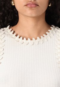 Top de punto blancoso con acentos texturizados en forma de hoja a lo largo del escote. Presenta un collar redondeado y una superficie de tejido suave.