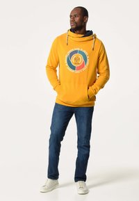 Un sweatshirt jaune avec un imprimé graphique présentant des icônes du tennis, une capuche avec cordon de serrage et des poches avant, associé à un jean foncé et des chaussures blanches.