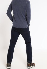 Mann steht mit den Händen in den Taschen, trägt einen strukturierten dunkelblauen Pullover, dunkle Jeans und sneakers mit camouflagemuster, schaut auf einen weißen Hintergrund.