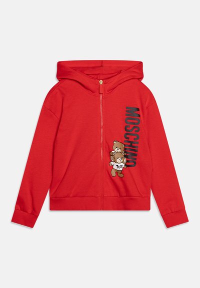 Rød hettegenser med glidelås laget av mykt stoff, med et bjørnesymbol og "Moschino" i fet, svarte bokstaver trykt ved siden av. Hetter og ribbestrikkede mansjetter.