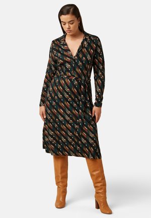 Vrouw draagt een lange mouwen, knielange jurk met diagonale patronen in bruin, groen en zwart en beige kniehoge laarzen.