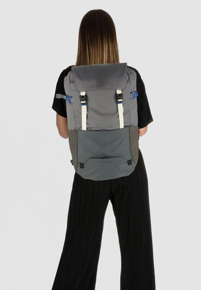 Mochila gris con un diseño estructurado, con herrajes azules y negros, correas ajustables y un acabado texturizado. Logo visible en la parte inferior.
