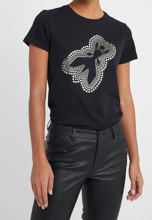 Mujer con una camiseta negra de manga corta con un patrón de tachuelas metálicas en forma de figura abstracta, combinada con pantalones de cuero negros.