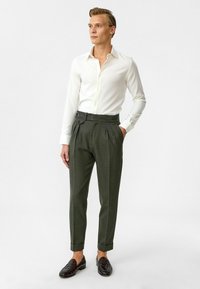 Witte langemouwen shirt, donker groene geplooide broek met zijgesp_rrng detail, en donkere geweven schoenen, staand tegen een effen achtergrond.