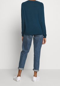 Maglietta a maniche lunghe teal realizzata in tessuto morbido, abbinata a jeans in denim blu con risvolto. Le sneakers bianche completano l'outfit.