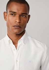 Camisa blanca de botones con collar tipo punta y un pequeño detalle bordado azul en el pecho. Textura suave, diseño ajustado.
