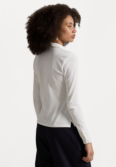 Long-sleeve white top made of lightweight fabric, featuring a collared neck, side slits, and button accents at the cuff.