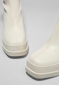 Valkoiset nahka nilkkurit, joissa on pyöreä kärki ja paksu kumipohja. Ominaisuuksiin kuuluvat näkyvät saumat ja sileä, teksturoitu pinta.