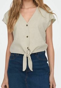 Světlé béžové tričko s krátkými rukávy, véčkovým výstřihem a předním zavazováním, zdobené hnědými knoflíky a mírně texturovanou látkou.